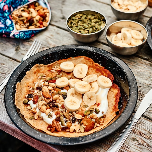 Une crêpe garnit avec des morceaux de bananes et plusieurs sortes de noix dans une assiette noire avec de petits pois blancs.