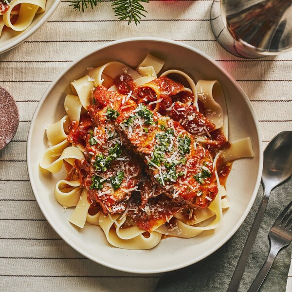 Au centre d’une nappe blanche lignée, il y a un grand bol de pappardelles avec une poitrine de dindon tranchée recouverte de sauce tomate, de gremolata et de parmesan. À droite, une fourchette et une cuillère, une bouteille et une coupe de vin rouge. À gauche, une bougie, un petit bol de gremolata et du sapinage.