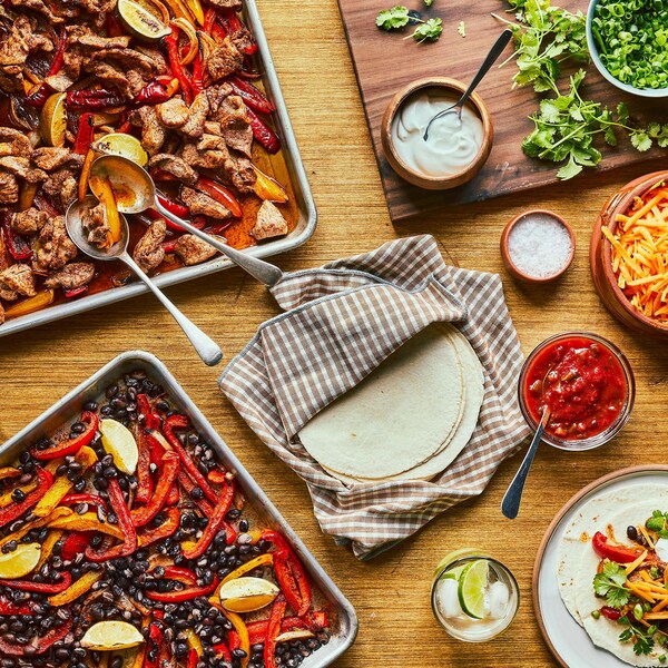 Une plaque de porc et de légumes ainsi qu’une plaque de haricots noirs et de légumes sont accompagnées d’une assiette de petites tortillas de blé, d’un bol de salsa, d’un bol de fromage, d’un bol de coriandre et d’assiettes avec des fajitas.