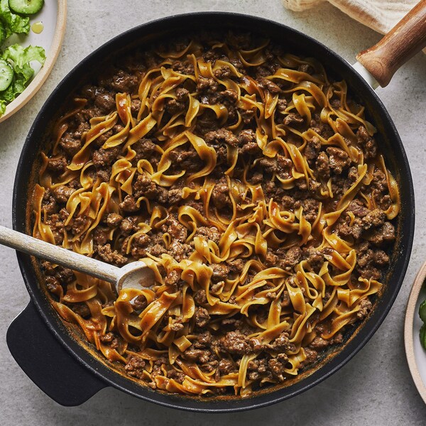 Une recette de Hamburger Helper maison dans une casserole avec une cuillère en bois, une main de sel et deux bols de salade verte.