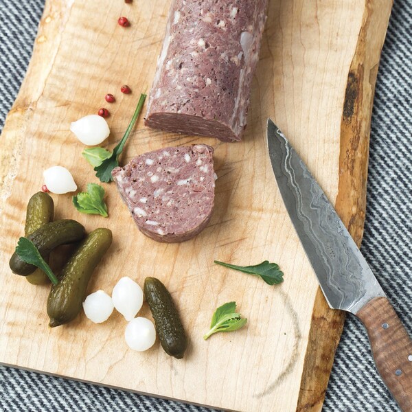 Un morceau de terrine de cerf sur une planche à découper. La terrine est accompagnée de petits cornichons et d'oignons marinés.