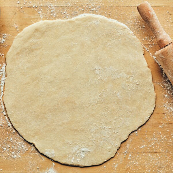 Sur un comptoir, est déposé la pâte à tarte, un rouleau pour abaisser la pâte et une assiette. 