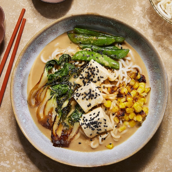 Un bol rempli de soupe ramen crémeuse garnie avec des bok boys, des pois mange-tout, du tofu et du maïs.