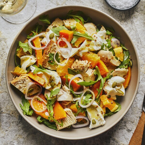 Dans un bol on retrouve un mélange coloré de dindon, de courge, de burrata, d'échalotes françaises et d’oranges. À droite, un pain grillé dans une assiette avec une coupe de vin blanc, et à gauche, des ustentiles et une seconde assiette de pain grillé.