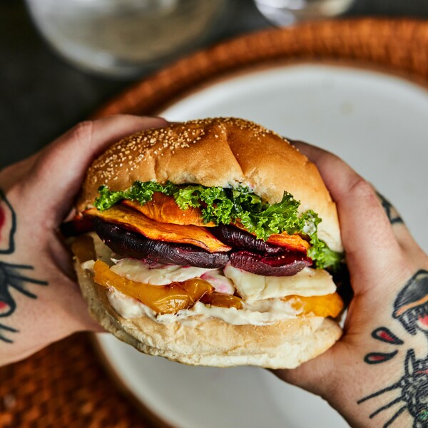 Il y a deux mains qui tiennent le sandwich et en arrière plan, il est possible de voir une assiette sur un napperon. 