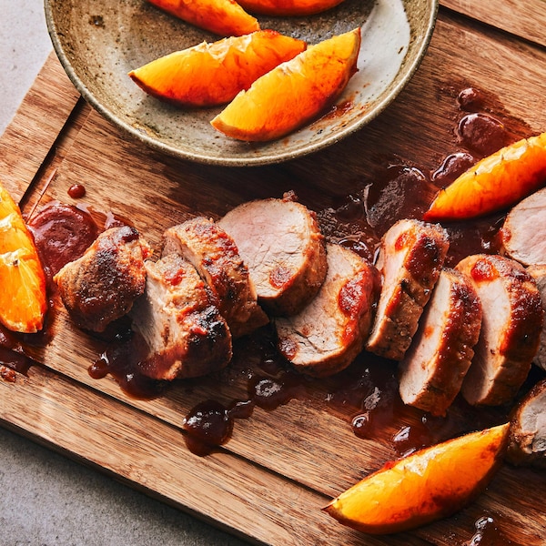 Sur une planche de bois, un filet de porc est coupé en tranches avec des quartiers d’orange et un filet de sauce au piment chipotle.
