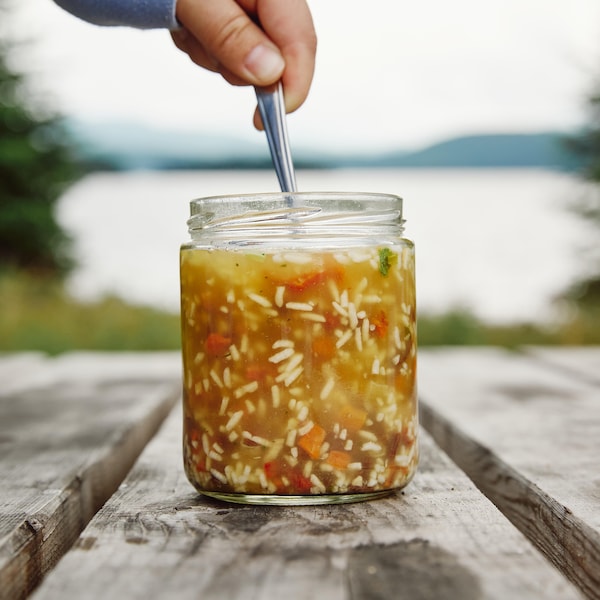 Un pot en verre, contenant de la soupe au jerky et au riz, est déposé sur une table en bois. Un main est au-dessus du pot en verre et elle tient la cuillère qui est dans le pot.