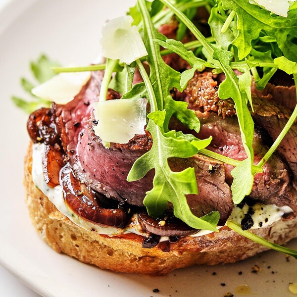 Une tranche de pain garnie de tranches de steak, d'oignons caramélises, de copeaux de fromage et de roquette.