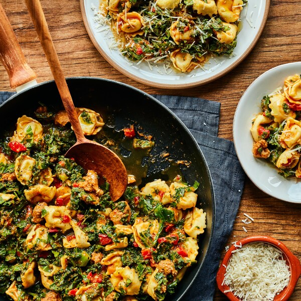 Sur une table est déposé trois assiettes et une poêle, tous contenant les tortellinis. Il y a aussi un petit bol de fromage parmesan et des ustensiles à çôté des assiettes. 