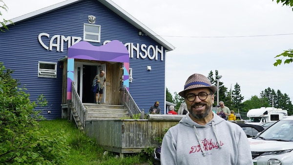 Le directeur du Festival en Chanson porte un chapeau et sourit à l'objectif devant un bâtiment du camp en chanson.