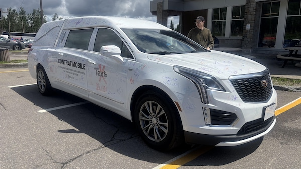 Un corbillard blanc de marque Cadillac est garé dans un stationnement asphalté. Sa carrosserie est recouverte de nombreuses signatures et messages manuscrits colorés, ainsi que d'une inscription textuelle promotionnelle ou de sensibilisation sur le flanc.