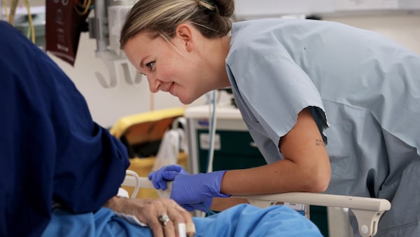 Une infirmière en tunique bleue est penchée au-dessus d'une personne couchée dans un lit et sourit.