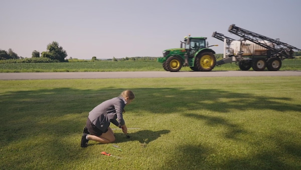 Une chercheuse récolte un échantillon de sol alors que passe un tracteur avec un pulvérisateur de pesticides.