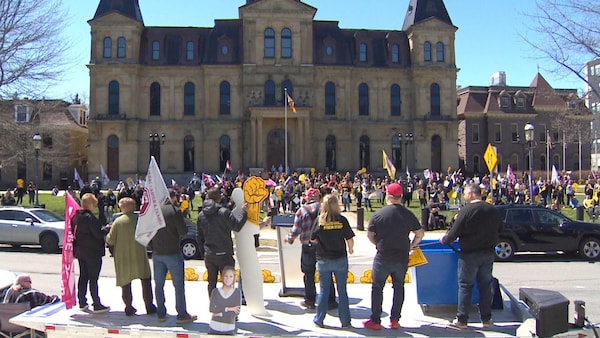 Une foule de quelques centaines de personnes avec des pancartes et des drapeaux, devant l'édifice de l'Assemblée législative, un jour de printemps ensoleillé.