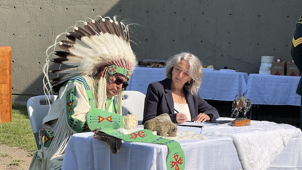 Un homme portant une coiffe traditionnelle autochtone signe un document en compagnie d'une femme, assis devant une table.
