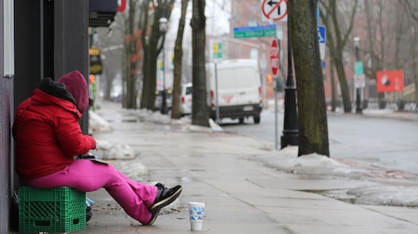 Une femme habillée en rose, un capuchon sur la tête, est assise sur une caisse de lait avec un gobelet devant elle, sur le trottoir d'une rue déserte à la fin de l'hiver.