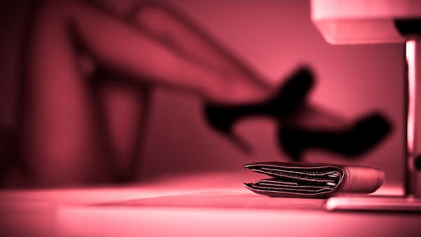 Un portefeuille posé sur une table de chevet. En arrière-plan, les jambes d'une femme en talons hauts, couchée, dans une lueur rose.