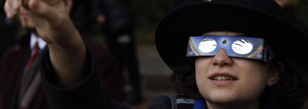 Un enfant portant des lunettes pour l'éclipse solaire regarde vers le ciel et pointe son doigt dans cette direction.