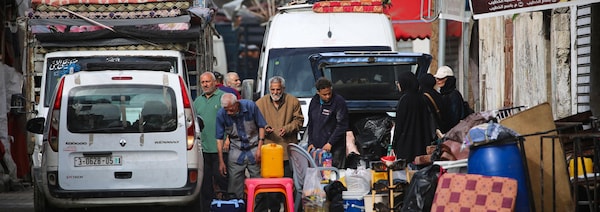 Une dizaine d'adultes sont debout dans une rue, à côté de sacs, de tables et de chaises de plastique ainsi que de coussins en attente d'être chargés dans un véhicule.