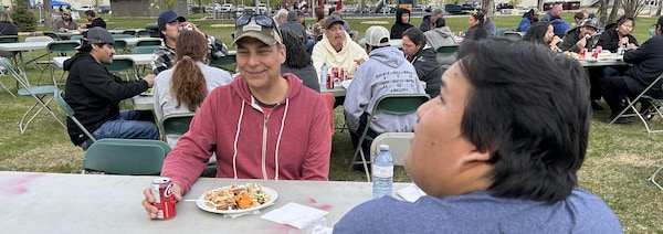 Une vingtaine de personnes mangent et jasent autour de tables communautaires à l'extérieur.