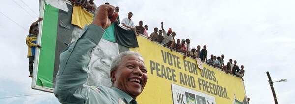Souriant et le poing levé, Nelson Mandela se tient devant un panneau sur lequel se tiennent de nombreuses personnes.