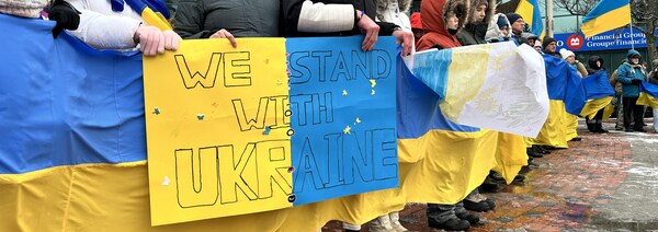 Des dizaines de personnes alignées dehors en hiver dans le froid tiennent une longue bannière jaune et bleu et une affiche avec le message « We Stand With Ukraine ».