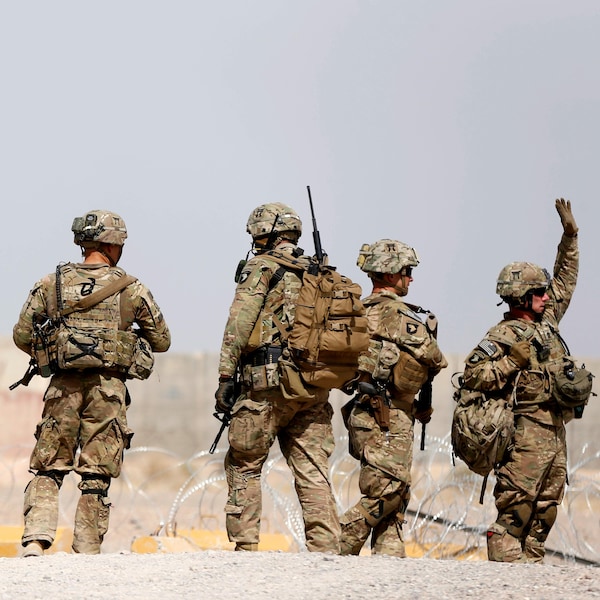 Six soldats américains marchent sur un sol terreux, le long d'une clôture de barbelés.