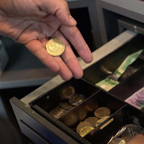Une main, se trouvant au-dessus d'une caisse enregistreuse contenant des billets d'argent canadien et de la monnaie canadienne, tient une pièce de monnaie dorée.