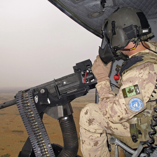 Un soldat canadien est assis dans un hélicoptère, devant un fusil mitrailleur. 
