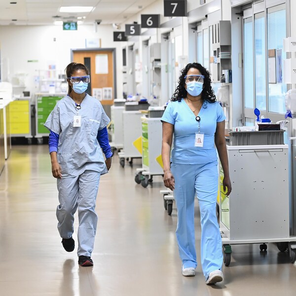 Deux infirmières avec masque et lunettes de protection marchant dans un couloir d'hôpital.