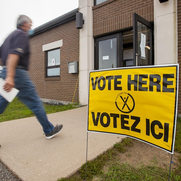 Un homme marche à grands pas vers la porte d'entrée d'un édifice à côté duquel est plantée une affiche jaune qui dit «Votez ici».