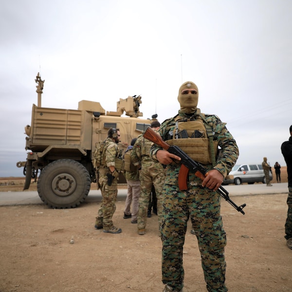 Un soldat masqué porte une arme au premier plan. Au second est posté un véhicule blindé.