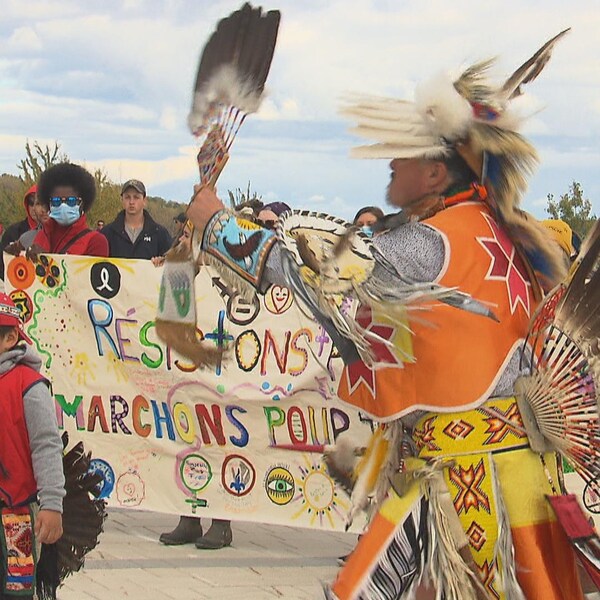 Un homme et un garçon, en habits traditionnels autochtones, dansent et des personnes rassemblées assistent à la prestation.