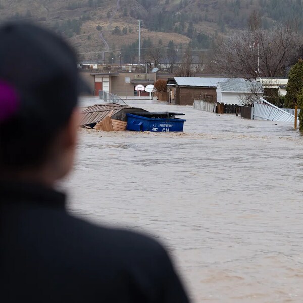 Une femme est debout devant un secteur inondé de Merritt qui comprend quelques maisons et des clôtures renversées.