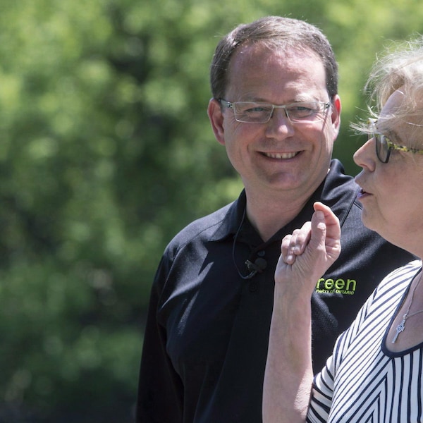 Mike Schreiner, portant un t-shirt noir, regarde Elizabeth May, vêtue d'une robe.