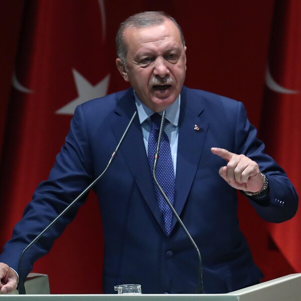 Le président Erdogan, debout, parle dans des micros, devant des drapeaux turcs.