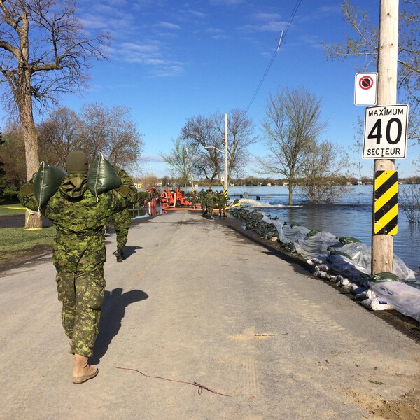 Un soldat des Forces armées canadiennes apporte des sacs de sable devant servir à renforcer une digue dans le secteur du boulevard Lalande et de la rue Rose, dans Pierrefonds-Roxboro, pendant que d'autres soldats travaillent au loin.