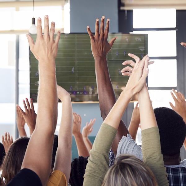 Plusieurs personnes de dos, les mains en l'air, célèbrent un jeu de football à la télévision.