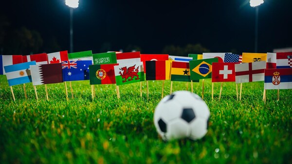 Un ballon de soccer est posé devant une multitude de drapeaux internationaux piqués sur une pelouse.