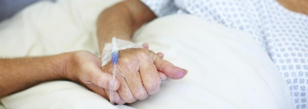 Gros plan sur une personne alitée aux soins palliatifs. Un individu la tient par la main. 