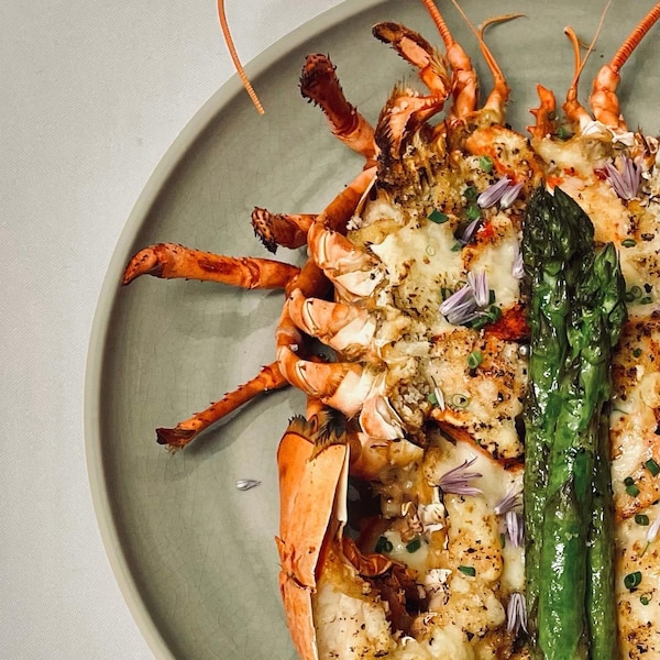 Dans une assiette repose un homard cuisiné avec des asperges et différentes herbes. 