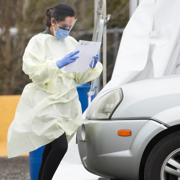 Une membre du personnel médical vêtue d'une jaquette et d'un masque lit un document devant une voiture à un centre de dépistage du coronavirus au volant.