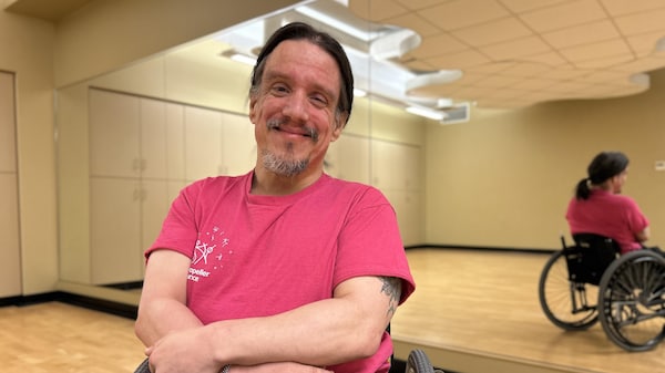 Un homme en fauteuil roulant sourit dans un studio de danse. Son reflet apparaît dans le miroir derrière lui.