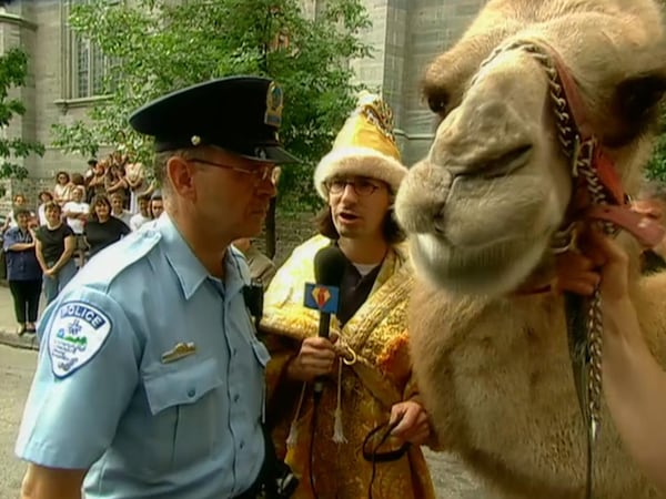 Jean-René Dufort déguisé en roi mage tient un chameau et un micro devant un policier de la ville de Montréal.
