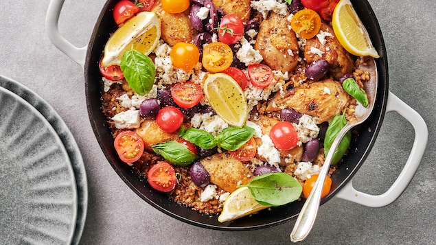 Une casserole remplie de pilons de poulet, de riz brun, de tomates, d'olives, de fromage et de basilic.