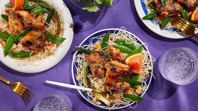 Des hauts de cuisses de poulet servis avec des vermicelles de riz, des oignons verts cuits, des pois sucrés et un quartier d'orange.