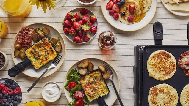 Sur une grande tablée, il y a deux assiettes avec une coupelle à raclette remplie d’une omelette, des patates grillées et de la pancetta cuite. Il y a aussi un bol de fraises, une assiette de pancakes avec des petits fruits, un bol de nutella maison, un bol de yogourt et un pichet de jus d’orange. En bas à droite, il y a des pancakes qui cuisent sur le poêle à raclette.