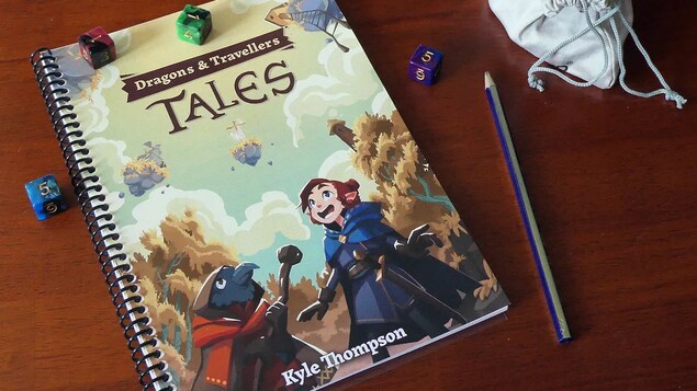 Le livret du jeu Dragons & Travellers' Tales est déposé sur une table avec un crayon et des dés.