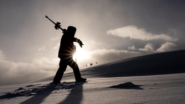 Un homme marche de profil dans la neige devant le soleil avec ses skis dans ses bras. 