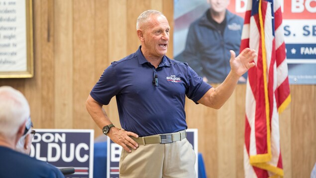 Don Bolduc, qui porte un polo bleu, parle à une foule, avec un drapeau américain derrière lui.